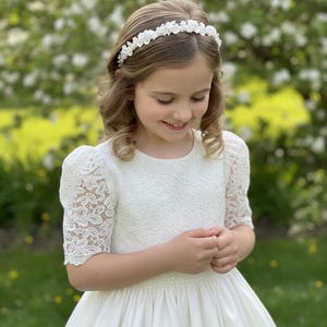 May include: A young girl wearing a white dress with lace sleeves and a satin skirt. She is also wearing a floral headband. The background is a blurred view of a garden with white flowers.