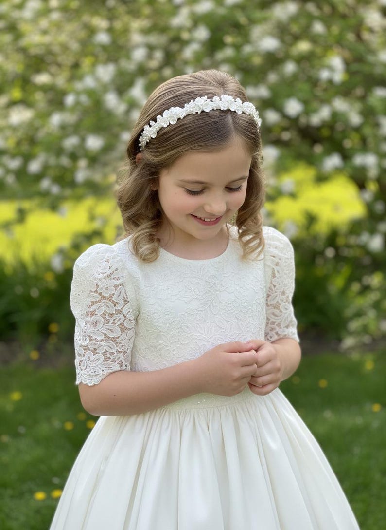May include: A young girl wearing a white dress with lace sleeves and a satin skirt. She is also wearing a floral headband. The background is a blurred view of a garden with white flowers.