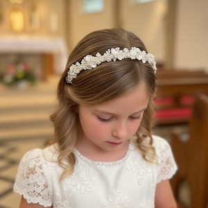 May include: A young girl wearing a white dress and a floral headband with pearls and crystals. The headband is centered on her head, and she is holding a small white book. The background is a church.