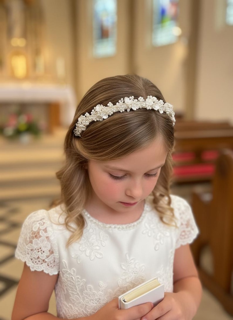 May include: A young girl wearing a white dress and a floral headband with pearls and crystals. The headband is centered on her head, and she is holding a small white book. The background is a church.