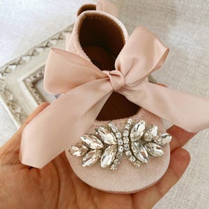 Peut inclure: Chaussures de b&eacute;b&eacute; en velours rose avec un grand embellissement en strass et un n&oelig;ud en satin assorti.
