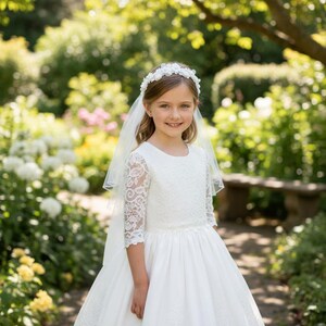 May include: A young girl in a white communion dress with lace sleeves and a matching floral headband. She is wearing a sheer veil and standing in a garden with a stone bench and lush greenery.