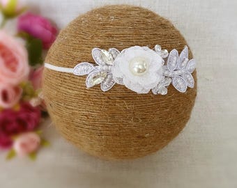 Könnte beinhalten: Ein weißes Stirnband mit einer Perlen- und Spitzenblume. Das Stirnband befindet sich auf einem braunen Hintergrund.