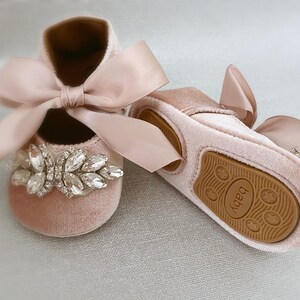 Peut inclure: Une paire de chaussures pour b&eacute;b&eacute; avec une tige en velours rose, un grand embellissement en strass et un n&oelig;ud en satin. Les chaussures ont une semelle en caoutchouc marron avec le mot "Baby" imprim&eacute; dessus.