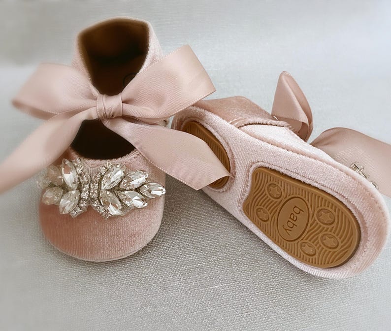 Peut inclure: Une paire de chaussures pour b&eacute;b&eacute; avec une tige en velours rose, un grand embellissement en strass et un n&oelig;ud en satin. Les chaussures ont une semelle en caoutchouc marron avec le mot "Baby" imprim&eacute; dessus.