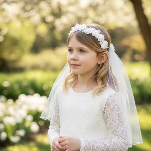 May include: A young person wearing a white lace dress with long sleeves, a floral headband, and a sheer veil. The dress has a simple, elegant design. The background is a blurred garden setting with white flowers.