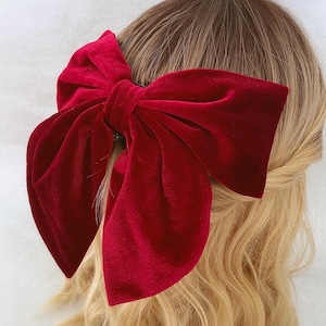 Könnte beinhalten: Eine große, rote Samt-Haarschleife. Die Schleife ist an einer Haarspange befestigt und wird im Haar getragen. Das Samtmaterial hat ein weiches, luxuriöses Aussehen. Die Schleife hat eine tiefe, satte rote Farbe.