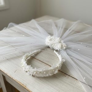 May include: A white bridal veil with a floral embellishment and a matching headband. The veil is made of sheer fabric and is decorated with small, sparkling accents. The headband features lace and pearl details.