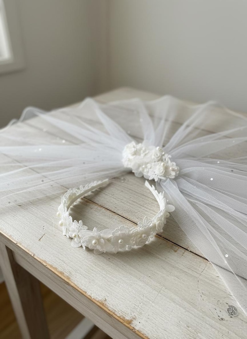 May include: A white bridal veil with a floral embellishment and a matching headband. The veil is made of sheer fabric and is decorated with small, sparkling accents. The headband features lace and pearl details.