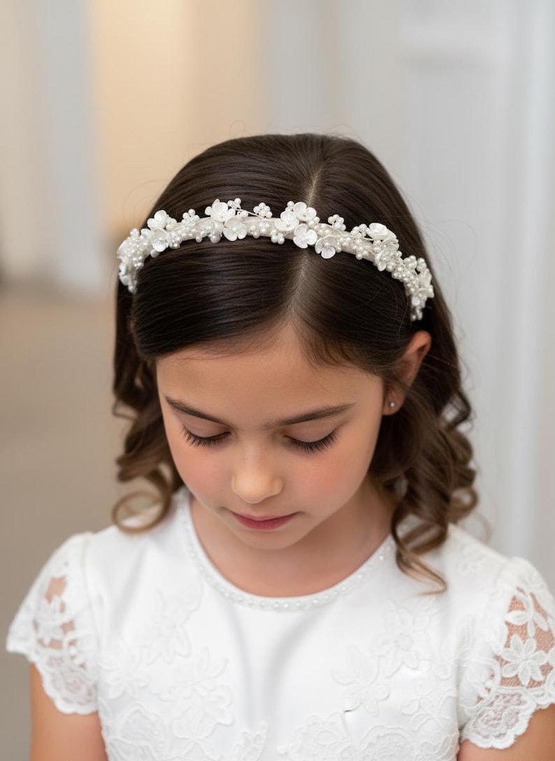 May include: A white floral headband with pearl accents adorns a young girl's dark hair. The headband features delicate white flowers and small pearls, creating an elegant and refined look. The girl is wearing a white dress.