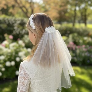 May include: A white lace headband and veil set. The headband features floral embellishments, and the veil is a sheer, delicate fabric. The person is wearing a white lace dress. The background is a blurred garden scene.