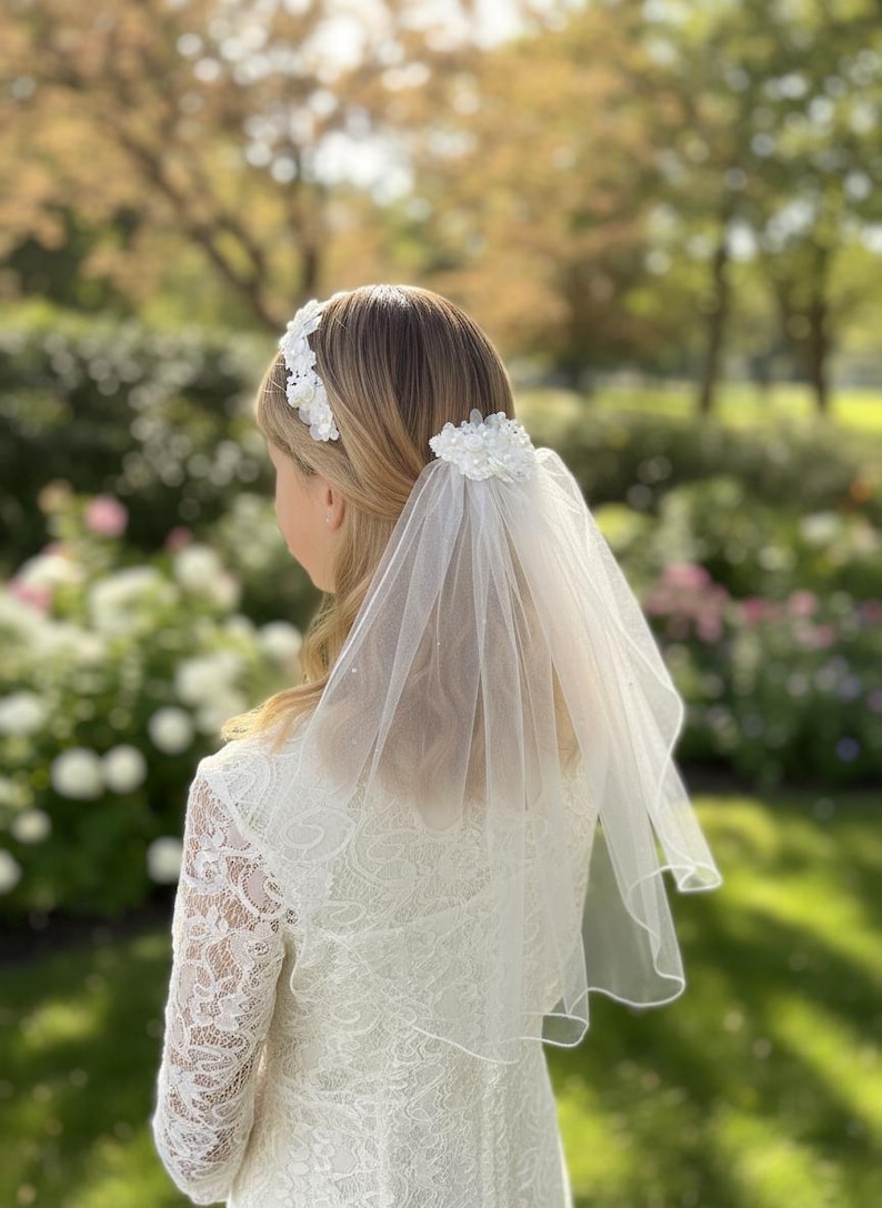 May include: A white lace headband and veil set. The headband features floral embellishments, and the veil is a sheer, delicate fabric. The person is wearing a white lace dress. The background is a blurred garden scene.