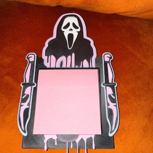 Puede incluir: Un organizador de escritorio negro y rosa con una máscara de Ghostface y dos cuchillos. El organizador tiene un bloc de notas rosa y detalles de pintura goteando. El diseño es un guiño a la franquicia de películas de terror Scream.