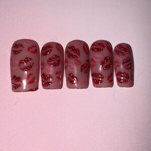 May include: Five artificial nails with a nude base, decorated with red glitter lips and pink lip outlines. The nails are displayed on a light pink surface, creating a Valentine's Day or romantic aesthetic.