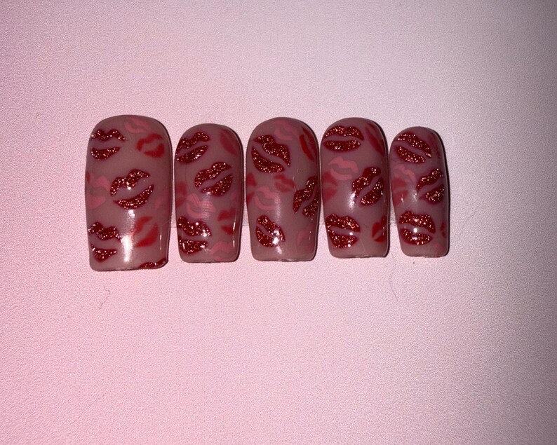 May include: Five artificial nails with a nude base, decorated with red glitter lips and pink lip outlines. The nails are displayed on a light pink surface, creating a Valentine's Day or romantic aesthetic.
