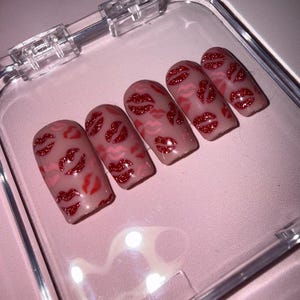May include: Five artificial nails with a nude base, decorated with red glitter lips. The nails are presented in a clear plastic case on a pink surface. The lip designs vary in size and placement.