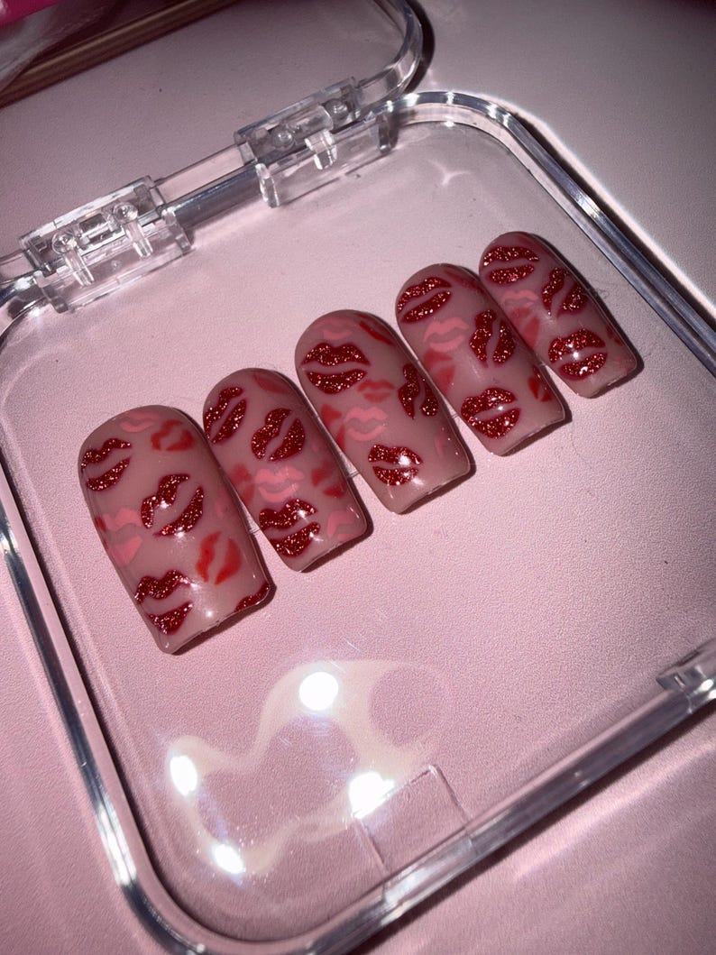 May include: Five artificial nails with a nude base, decorated with red glitter lips. The nails are presented in a clear plastic case on a pink surface. The lip designs vary in size and placement.
