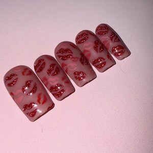 May include: Five artificial nails with a light pink base, decorated with red glitter lips. The lips vary in size and are scattered across the nail surface. The nails are arranged diagonally against a pink background.