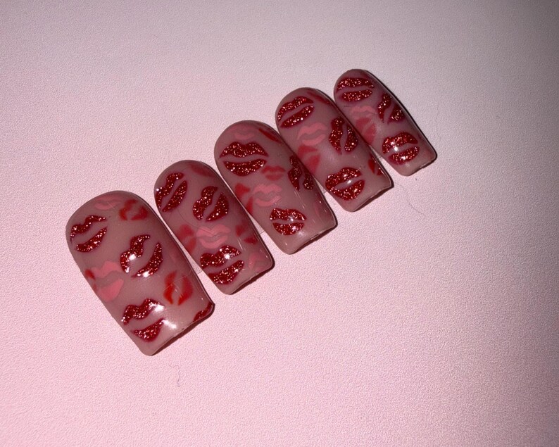 May include: Five artificial nails with a light pink base, decorated with red glitter lips. The lips vary in size and are scattered across the nail surface. The nails are arranged diagonally against a pink background.