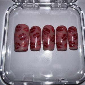 May include: Five artificial nails with a nude base, decorated with red glitter lips. The nails are presented in a clear plastic container, featuring a glossy finish and a playful design.