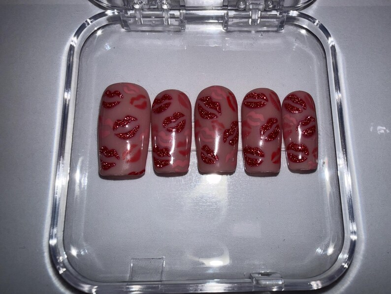 May include: Five artificial nails with a nude base, decorated with red glitter lips. The nails are presented in a clear plastic container, featuring a glossy finish and a playful design.