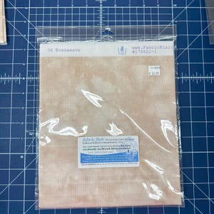May include: A package of 36 Evenweave fabric by Fabric Flair in a mottled tan color. The clear plastic packaging features a white label with the text 36 Evenweave, a hand wash icon, and a product label with care instructions and brand details.