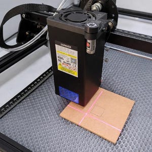 May include: A black laser engraving machine with a brown piece of material in the centre. The laser emits a pink light, creating a square pattern on the material. The machine is on a grey surface.