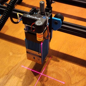 May include: A laser engraver with a blue and black frame, emitting a bright pink laser beam. The device has a "Danger" warning label and the text "Crossmate" is visible. The laser is focused on a wooden surface.