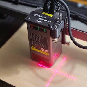 Könnte beinhalten: Eine silber-schwarze Lasergravurmaschine mit dem Text "Falcon 2 Crosshair - 22W" oben. Die Maschine emittiert zwei sich kreuzende rote Laserstrahlen. Das Gerät hat einen Ein-/Ausschalter und Kontrollleuchten.