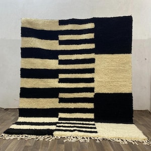 May include: A rectangular rug with a geometric pattern in black and cream. The rug's design includes stripes and blocks. The bottom edge has a fringe. The rug is made from a thick, woven material, suitable for a living space.
