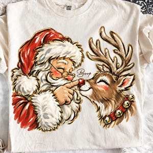 Könnte beinhalten: Ein cremefarbenes T-Shirt mit einer grafischen Illustration des Weihnachtsmanns, der die rote Nase eines Rentiers berührt. Das Rentier trägt ein rotes Halsband mit goldenen Glöckchen. Das Wort Boop steht in Schreibschrift zwischen den Figuren.
