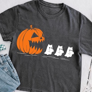 May include: A charcoal gray t-shirt featuring a large orange jack-o'-lantern pumpkin with a jagged mouth on the left side, appearing to chase three small white ghost figures lined up horizontally toward the right side of the garment.