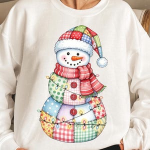 May include: White sweatshirt with a snowman design. The snowman has a patchwork hat, scarf, and body, with a carrot nose and button eyes. A string of lights wraps around the snowman's body, adding a festive touch for the winter season.