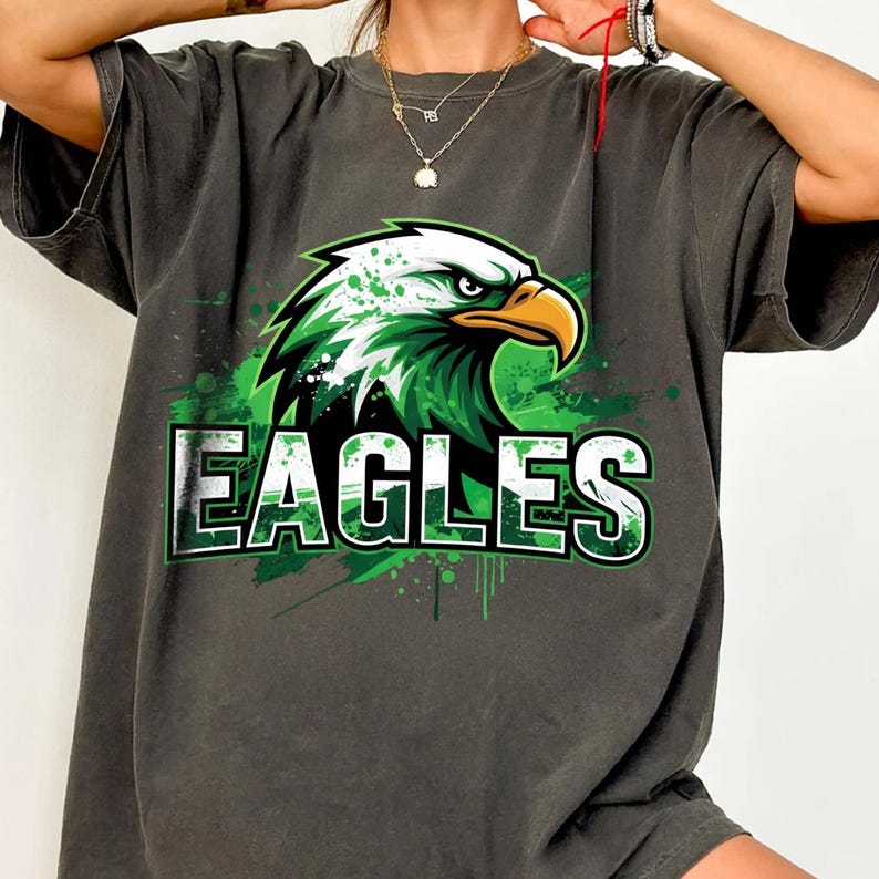 Op de afbeelding: Donkergrijs T-shirt met een groen-witte adelaarsafbeelding en het woord "EAGLES" in witte blokletters. Het adelaarsontwerp heeft groene verfspatten.