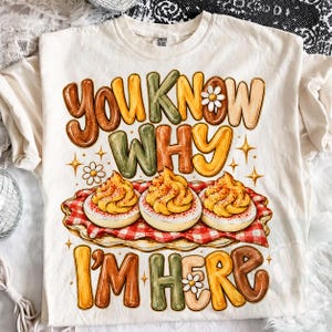May include: A cream-colored t-shirt featuring a graphic of three deviled eggs on a red and white checkered napkin. The text reads YOU KNOW WHY I'M HERE in large, multi-colored bubble letters, accented with small white daisy flowers and stars.