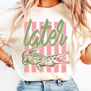 May include: Cream-colored t-shirt with a pink and white striped background. The shirt features the word "later" in green script above a detailed green and pink alligator illustration.