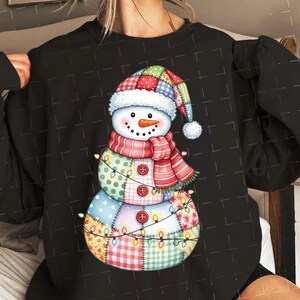 Puede incluir: Sudadera negra con un dise&ntilde;o de mu&ntilde;eco de nieve colorido. El mu&ntilde;eco de nieve tiene un cuerpo de retazos, una nariz de zanahoria y una bufanda a rayas rojas y blancas. Una guirnalda de luces envuelve al mu&ntilde;eco de nieve.