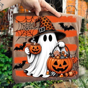 May include: A brown t-shirt featuring a graphic of a white ghost wearing an orange patterned witch hat. The ghost holds a small jack-o'-lantern mug and a large jack-o'-lantern bucket filled with sweets, surrounded by black bats and spiderwebs.