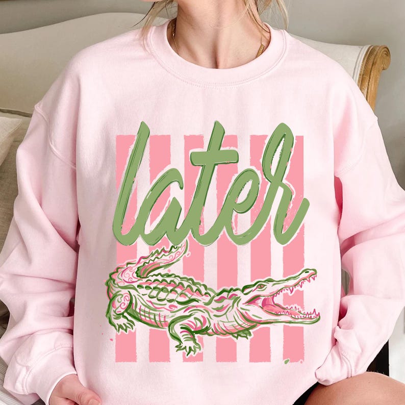 May include: A light pink sweatshirt featuring a green and pink alligator design with the word "later" in green script. The alligator is detailed with pink and green accents, set against a pink striped background.