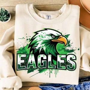 Op de afbeelding: Cr&egrave;mekleurige sweatshirt met een groene en zwarte adelaarsafbeelding met het woord "EAGLES" in een groen en wit lettertype. Het adelaarsontwerp heeft een spatteffect. De sweatshirt is gemaakt van een zacht materiaal.