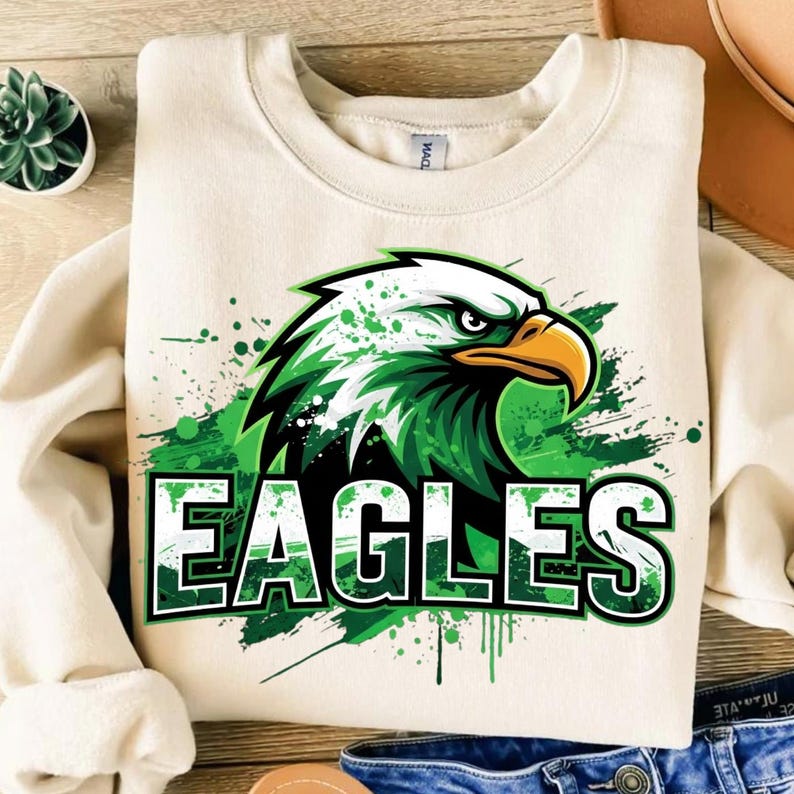 Op de afbeelding: Cr&egrave;mekleurige sweatshirt met een groene en zwarte adelaarsafbeelding met het woord "EAGLES" in een groen en wit lettertype. Het adelaarsontwerp heeft een spatteffect. De sweatshirt is gemaakt van een zacht materiaal.