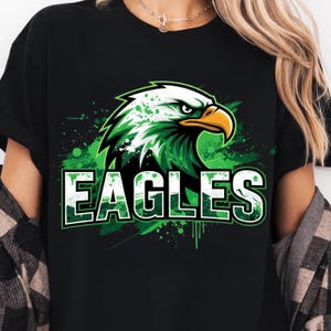 Op de afbeelding: Zwart T-shirt met een groen en witte adelaarsafbeelding en het woord "EAGLES". Het adelaarsontwerp heeft een groen en wit kleurenschema met een gele snavel. Het shirt heeft een ronde hals en korte mouwen.