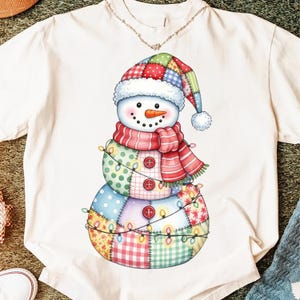 Puede incluir: Camiseta blanca con un dise&ntilde;o caprichoso de mu&ntilde;eco de nieve. El mu&ntilde;eco de nieve est&aacute; adornado con un gorro y bufanda de retazos, con una nariz de zanahoria y ojos de bot&oacute;n. Una guirnalda de luces envuelve el cuerpo de retazos.