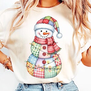 Puede incluir: Una camiseta color crema con un dise&ntilde;o caprichoso de mu&ntilde;eco de nieve. El mu&ntilde;eco de nieve est&aacute; adornado con un sombrero, bufanda y cuerpo de retazos, y est&aacute; decorado con una guirnalda de luces. El dise&ntilde;o incluye una nariz de zanahoria y botones.