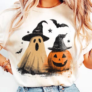 May include: Cream-colored t-shirt featuring a Halloween-themed graphic. The design includes a ghost wearing a witch hat, a jack-o'-lantern with a witch hat, bats, and stars. The ghost and pumpkin are orange and tan.