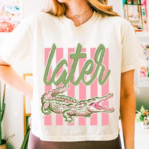 May include: Cream-colored t-shirt featuring a pink and white striped background with the word "later" in green script. Below the text is a detailed illustration of a green and pink alligator with its mouth open.