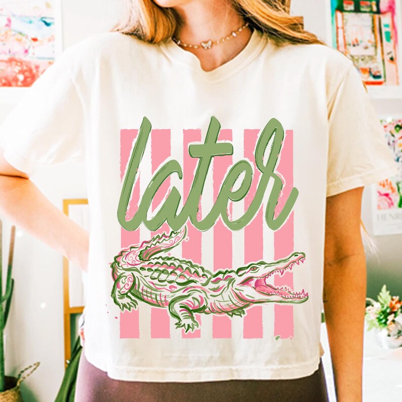 May include: Cream-colored t-shirt featuring a pink and white striped background with the word "later" in green script. Below the text is a detailed illustration of a green and pink alligator with its mouth open.