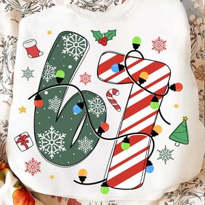 May include: White sweatshirt with a festive Christmas design. The number "67" is displayed, with the "6" in green with snowflakes and the "7" in red and white stripes, adorned with string lights and holiday icons.
