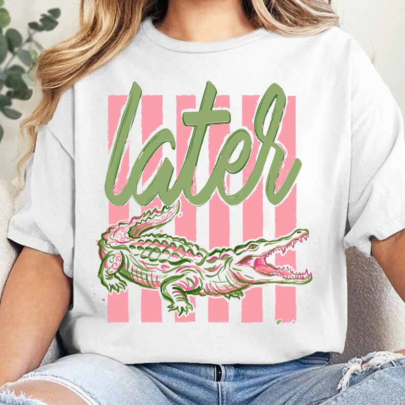 May include: White t-shirt with a pink and green alligator design. The word "later" is written in green script above the alligator. Pink vertical stripes are behind the text and alligator.