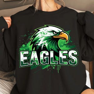 Op de afbeelding: Zwart sweatshirt met een groen en wit adelaarsontwerp en het woord "EAGLES". De afbeelding bevat groene verfspatten. De adelaar heeft een gele snavel en een geconcentreerde uitdrukking. Een sportief ontwerp.
